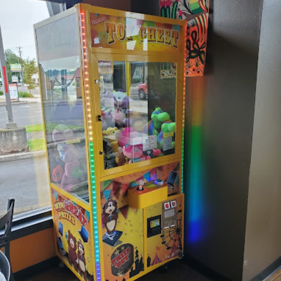 A yellow claw machine filled with plush toys stands inside Mami's Mexican Grill, reflecting the street outside.